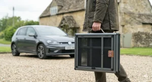 A person in a wax cotton jacket carrying a collapsed, portable folding small dog crate in a British village setting with a stone cottage background.